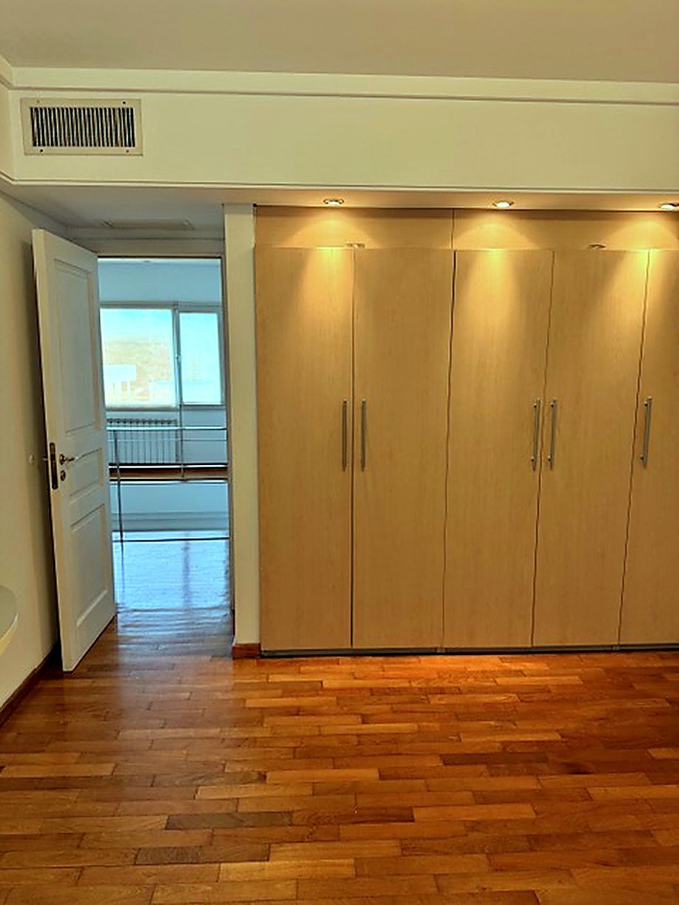 Bedroom with a full wall of light oak fitted wardrobes on oak parquet and adjoining room beyond